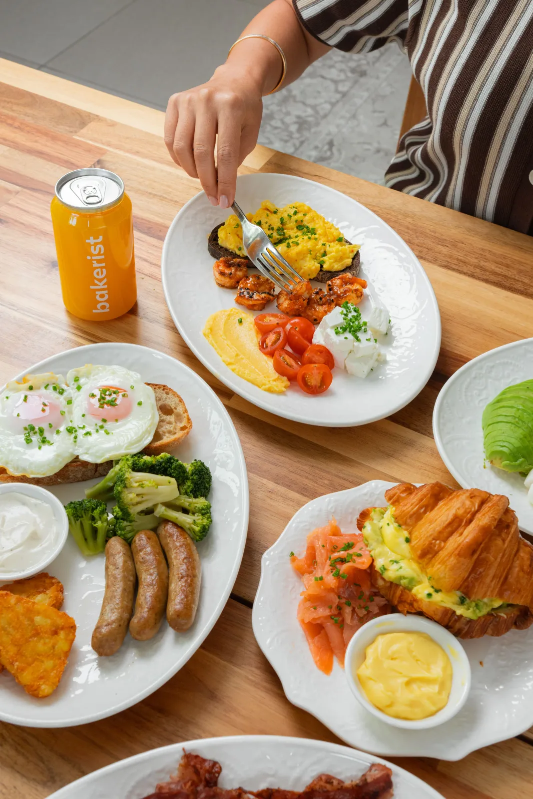 A breakfast table with eggs, sausages, hash browns, salmon croissant and a Bakerist juice can