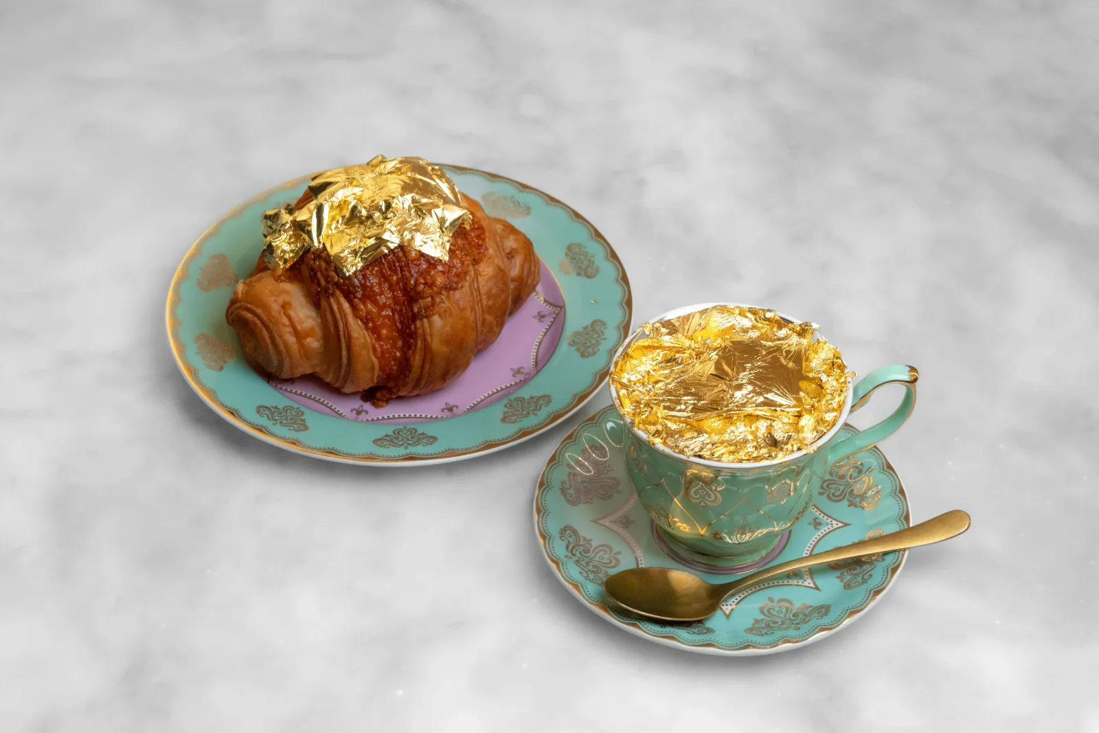 A croissant topped with edible gold leaf beside a gold-flecked coffee in a green and gold cup