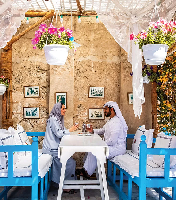 Two guests sharing Arabic coffee at a white table beneath hanging flowers, against a rustic sandstone wall