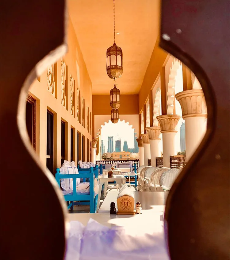 View through an ornate arch onto a sunlit terrace with blue benches and hanging lanterns