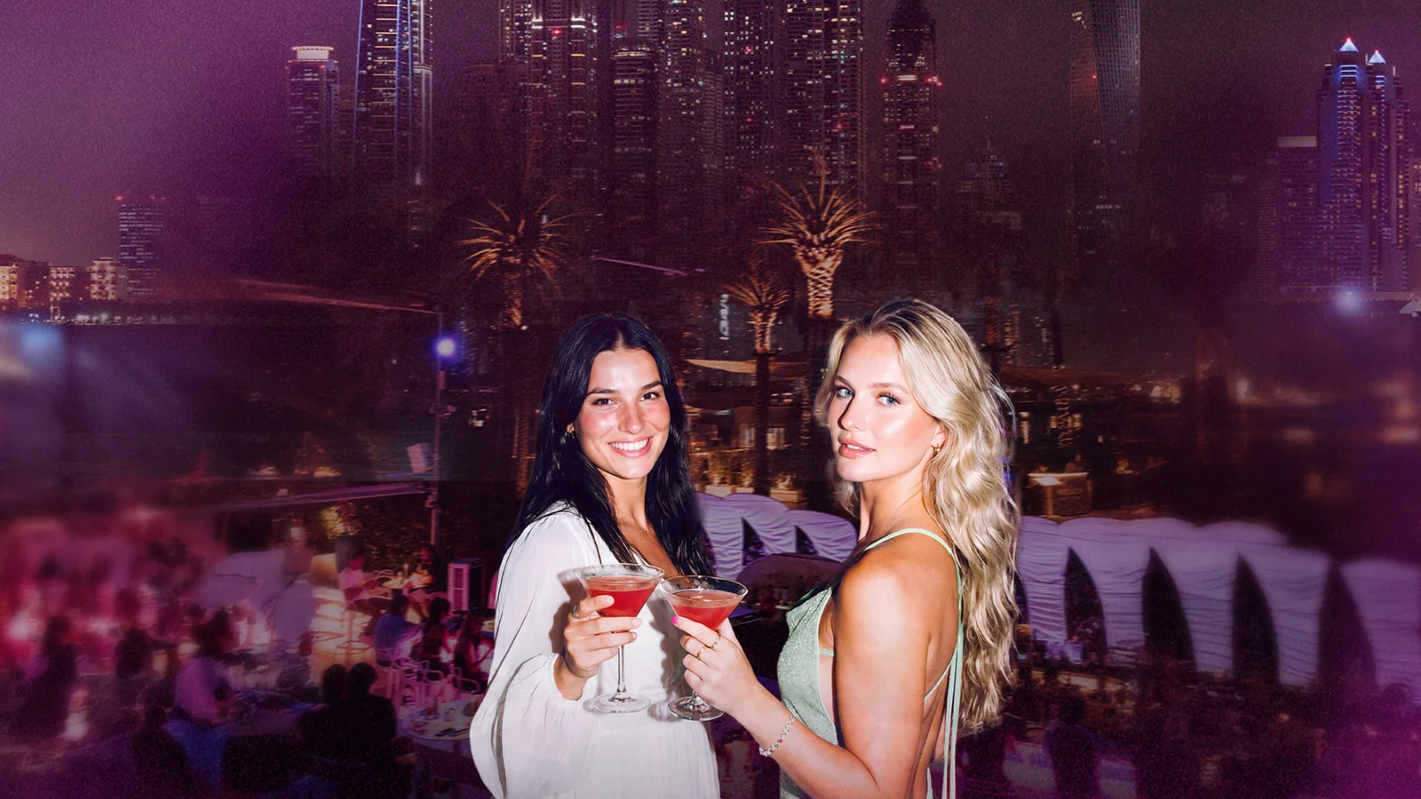 Two guests with cocktails at Praia at night, the Dubai skyline lit up behind them