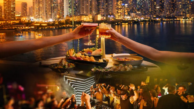 Hands toasting cocktails over a sushi boat and tacos under neon lounge lighting