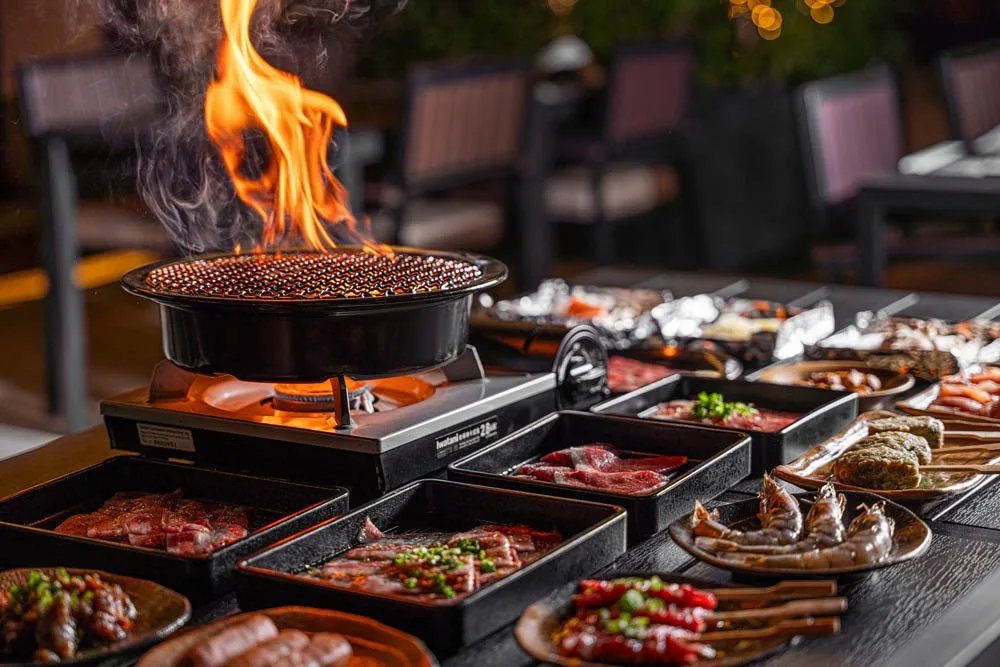 Charcoal grill flaming beside trays of marinated meat ready to cook