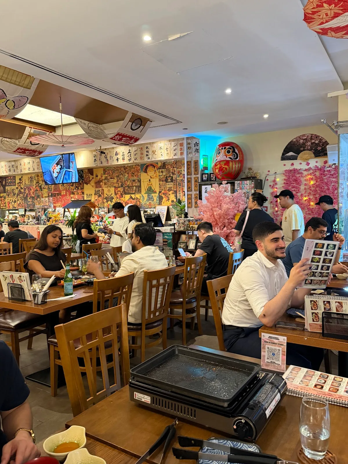 Busy Mogi-ya dining room with guests, a large red daruma and a cherry-blossom wall