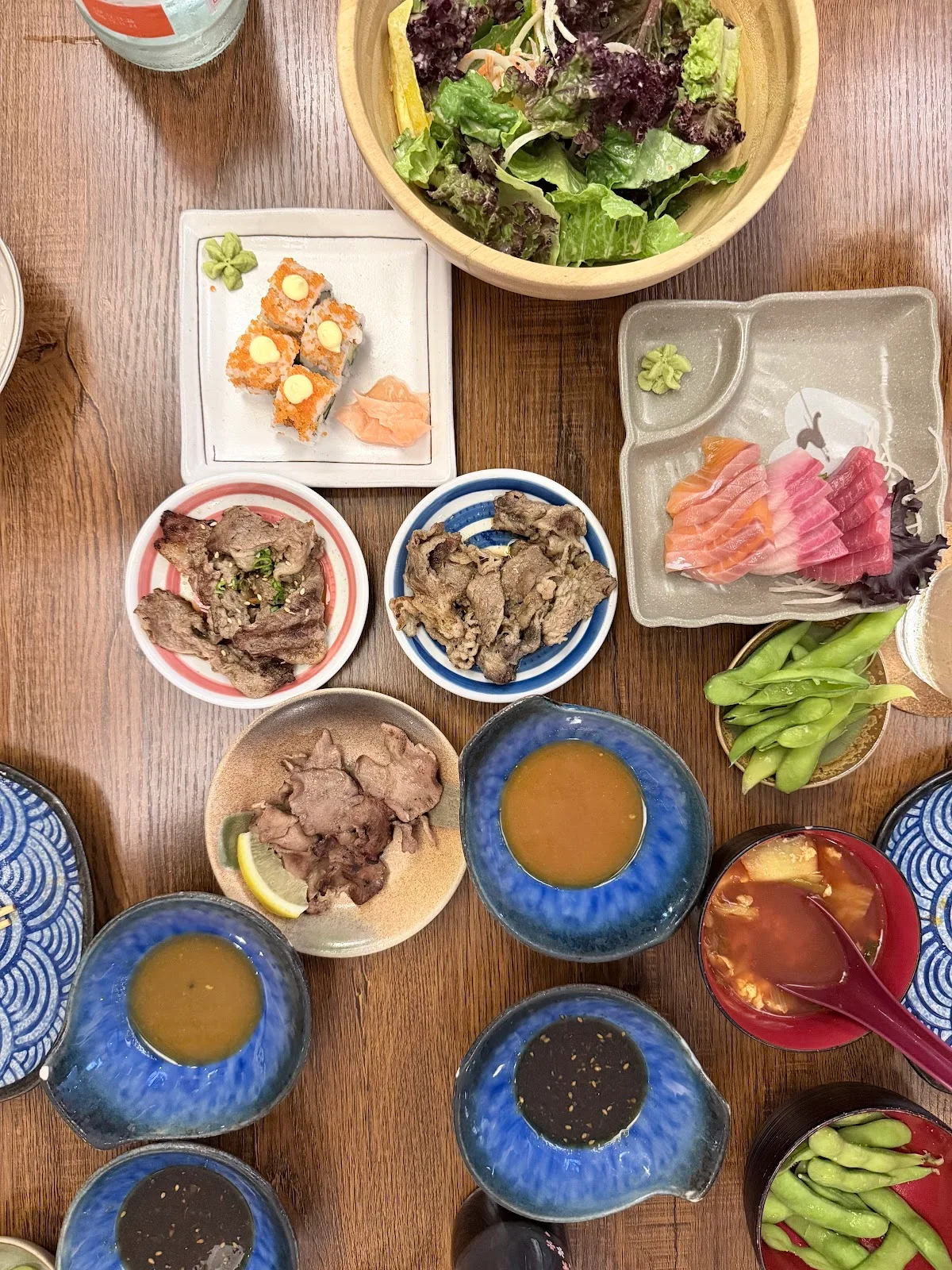 Overhead spread of sashimi, sushi, grilled beef plates and side dishes on a wooden table