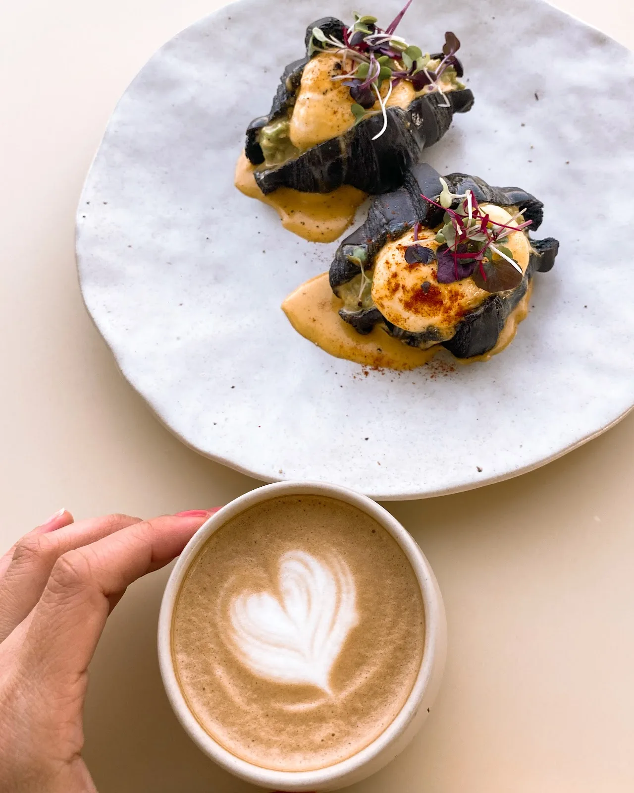 Charcoal eggs benedict topped with hollandaise and microgreens, beside a flat white.
