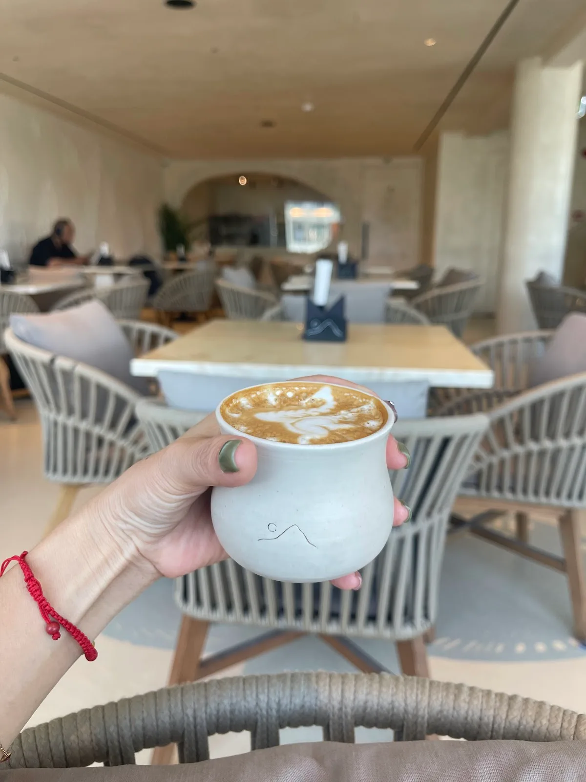 A flat white with latte art in a cream Lune cup, held in the bright lounge with rope chairs behind.