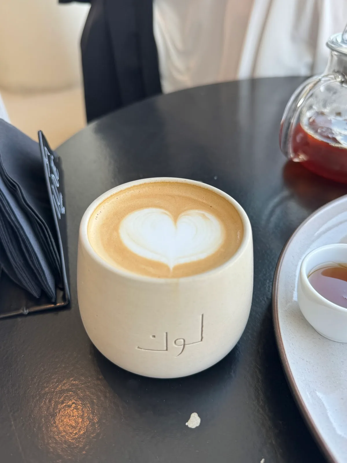 A flat white with heart latte art served in a cream Lune ceramic cup on a dark table.