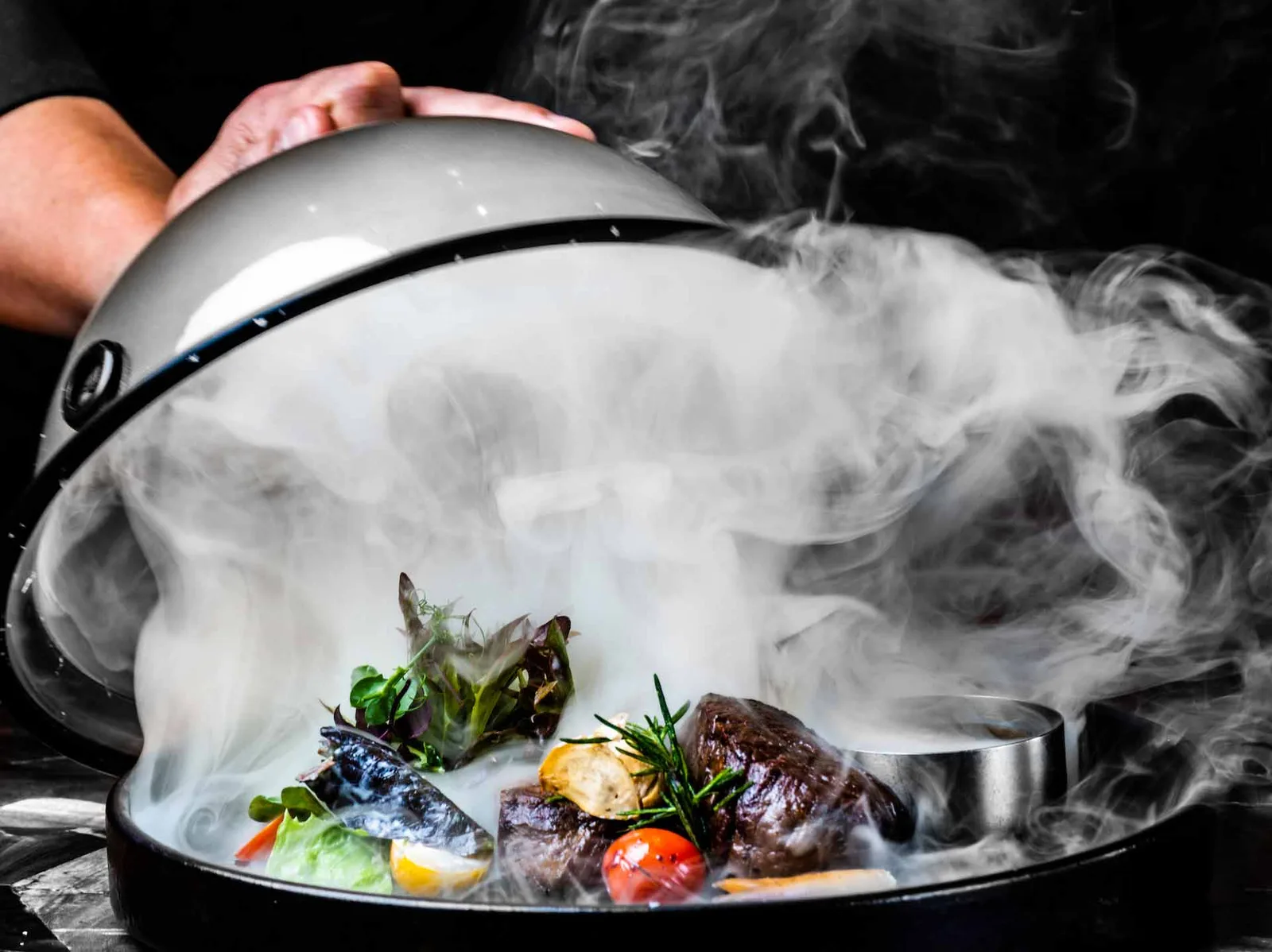 A chef lifting a smoke-filled cloche to reveal a smoked steak at Logs & Embers
