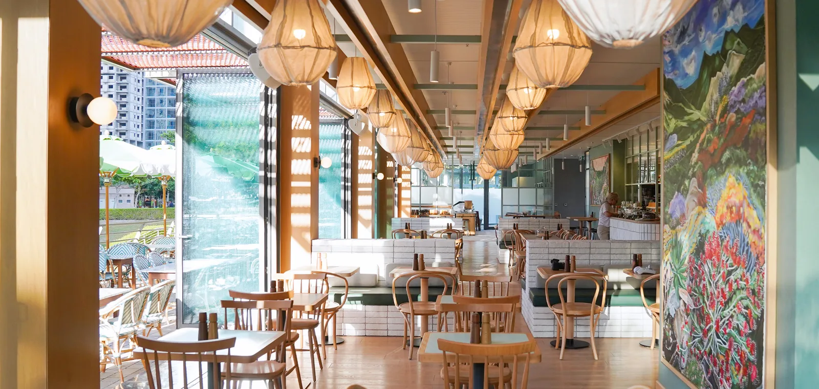 Sunlit dining hall with woven pendant lights and floor-to-ceiling glass opening to the park