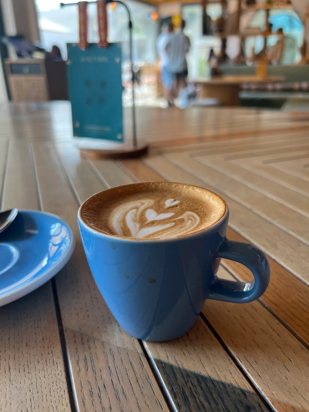 A flat white with leaf latte art in a blue cup on a sunlit wooden café table.