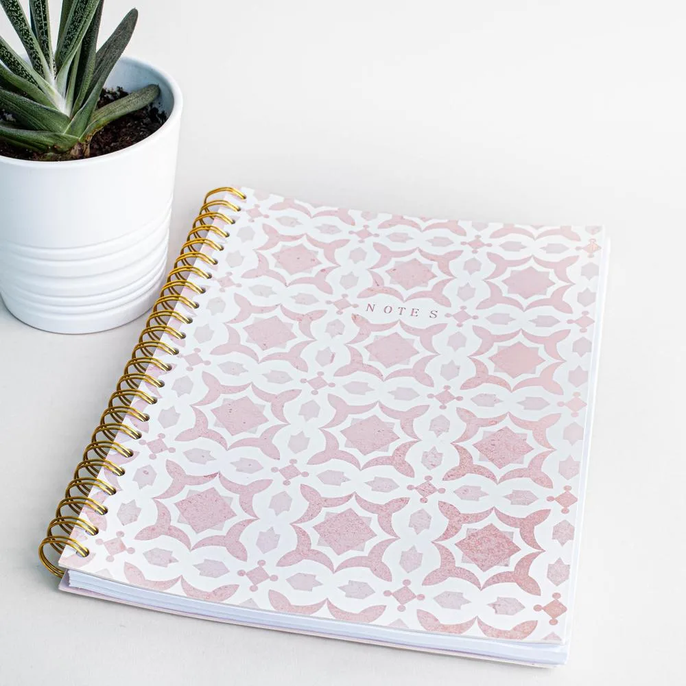 A spiral notebook with a pink Moorish-tile cover beside a succulent.