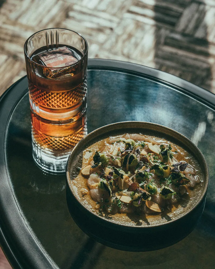 A delicate raw dish beside an amber cocktail