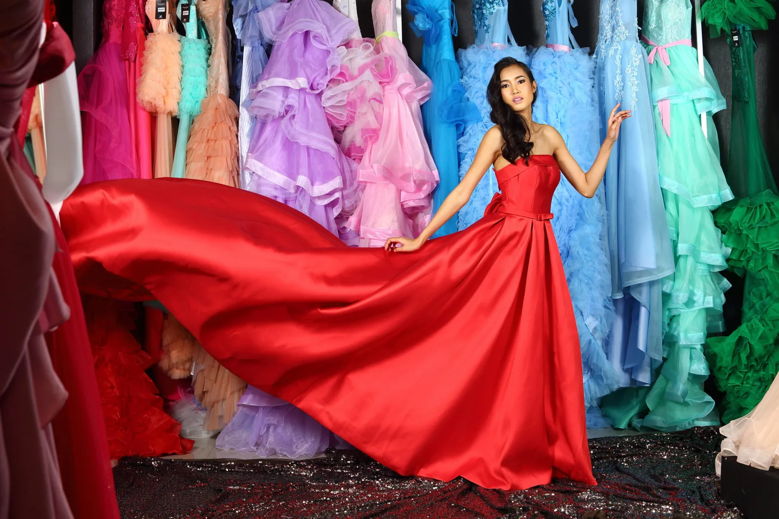 A model in a flowing red gown beside a rail of colourful occasion dresses