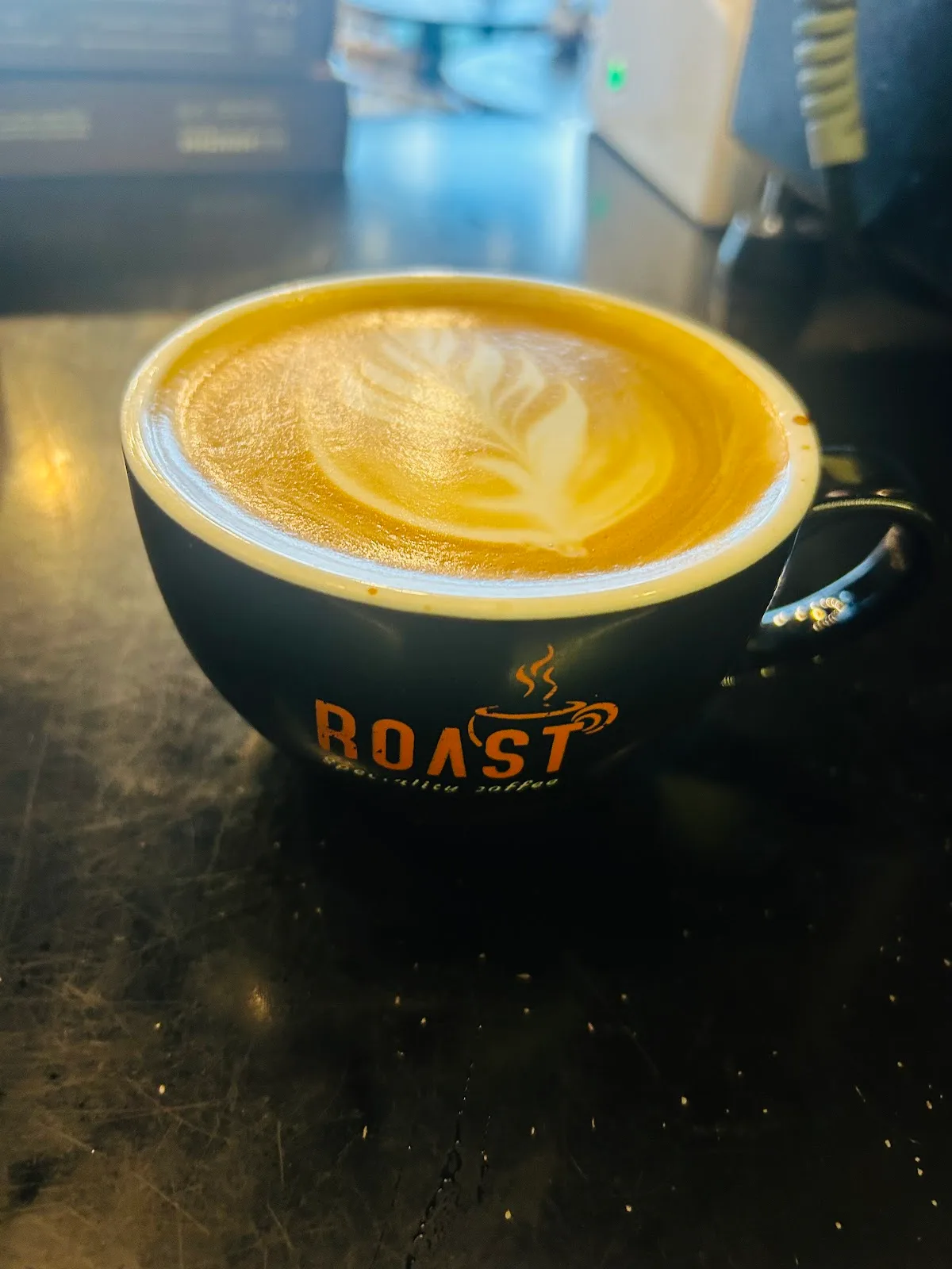 A Roast-branded cup of coffee with latte art on a dark counter
