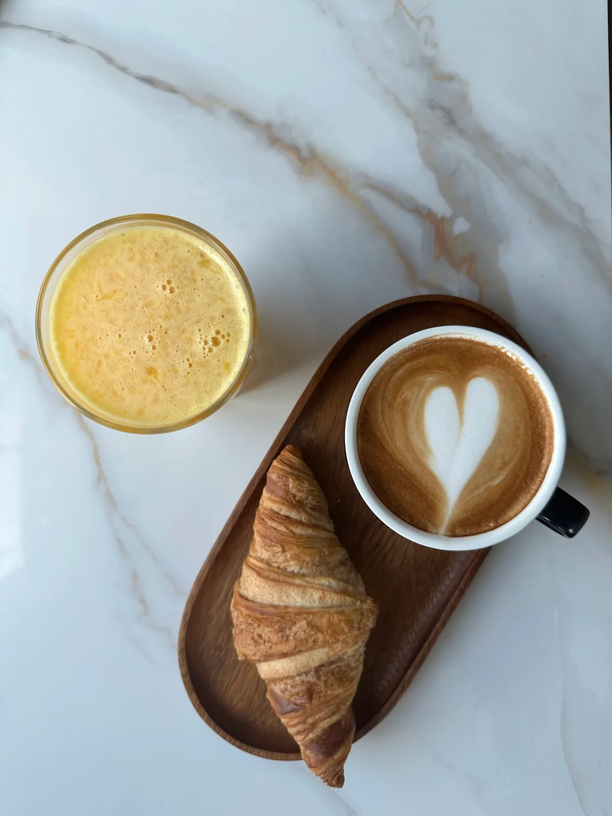 Latte with heart latte art, a croissant on a wooden board and fresh juice on a marble table