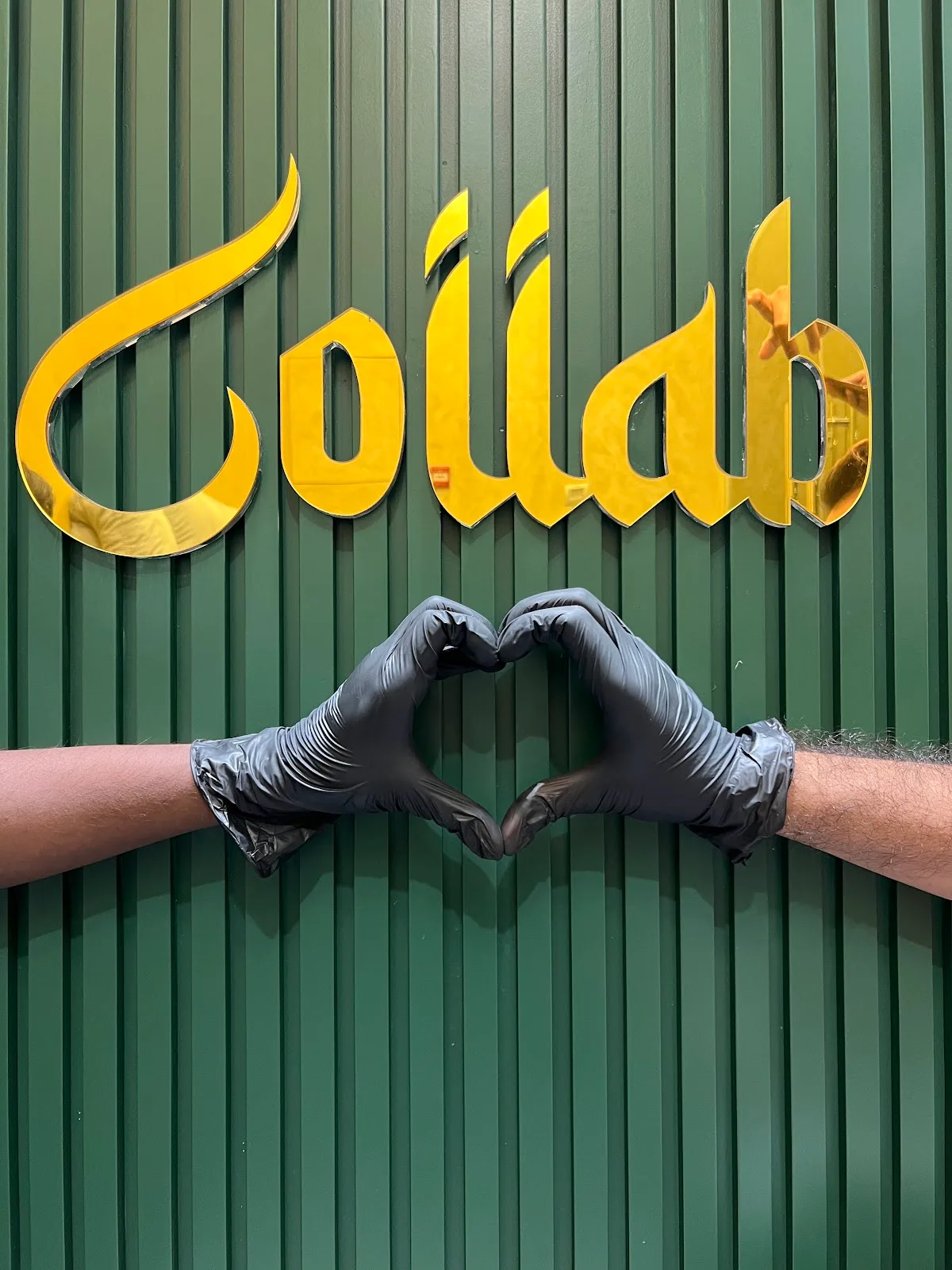 A team member making a heart shape with gloved hands beneath the gold Collab sign on a green wall