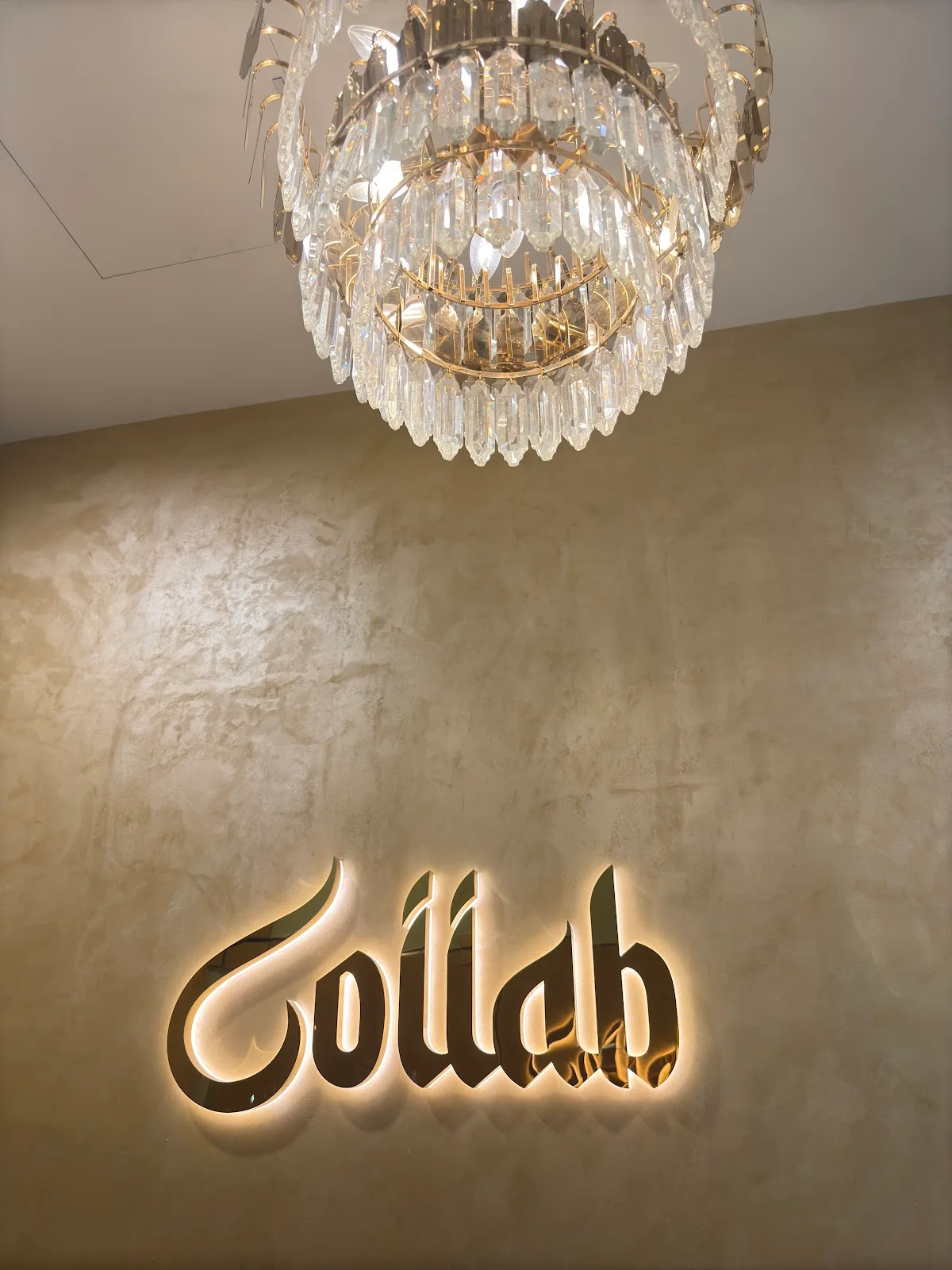 The backlit gold Collab sign glowing on a warm plaster wall beneath a crystal chandelier inside the studio