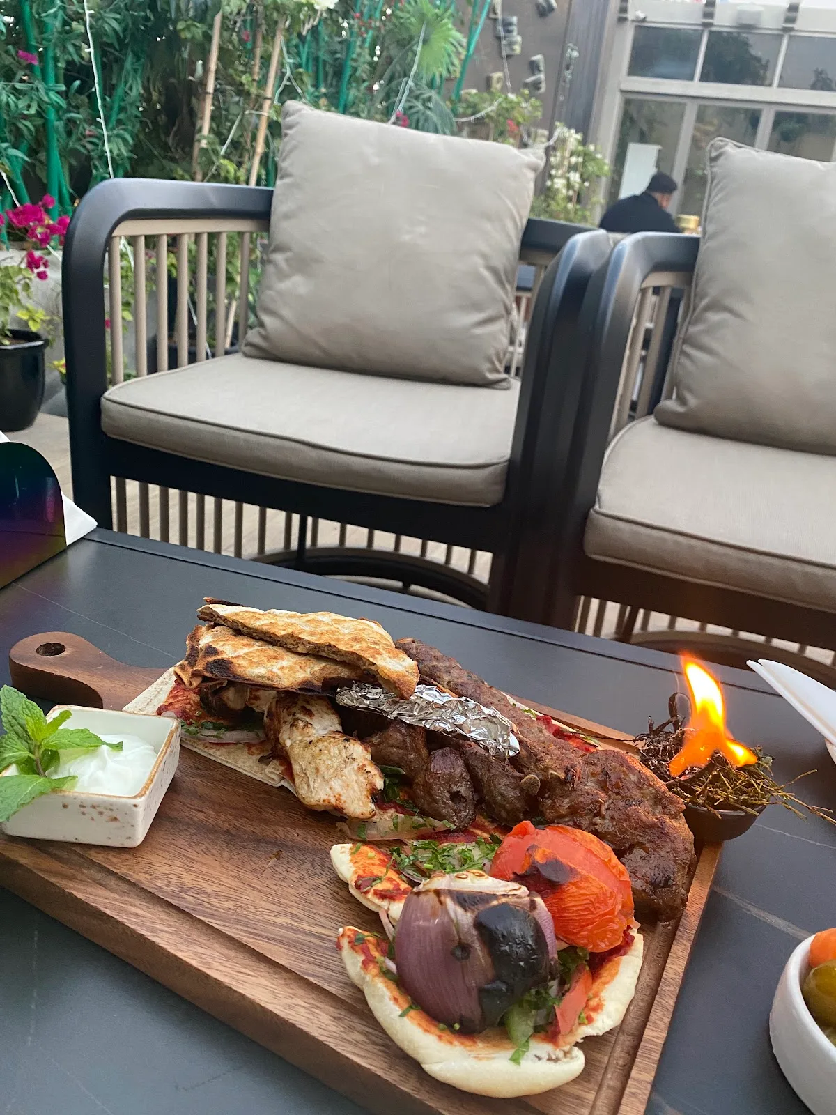 A mixed grill sharing board with grilled meats, flatbread and a small open flame, set on an outdoor table