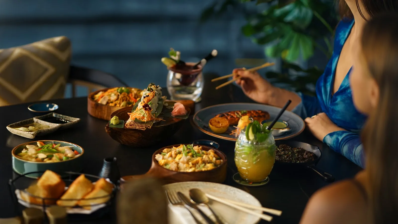 Guests sharing an array of dishes and a fresh cocktail across a dark table in the evening