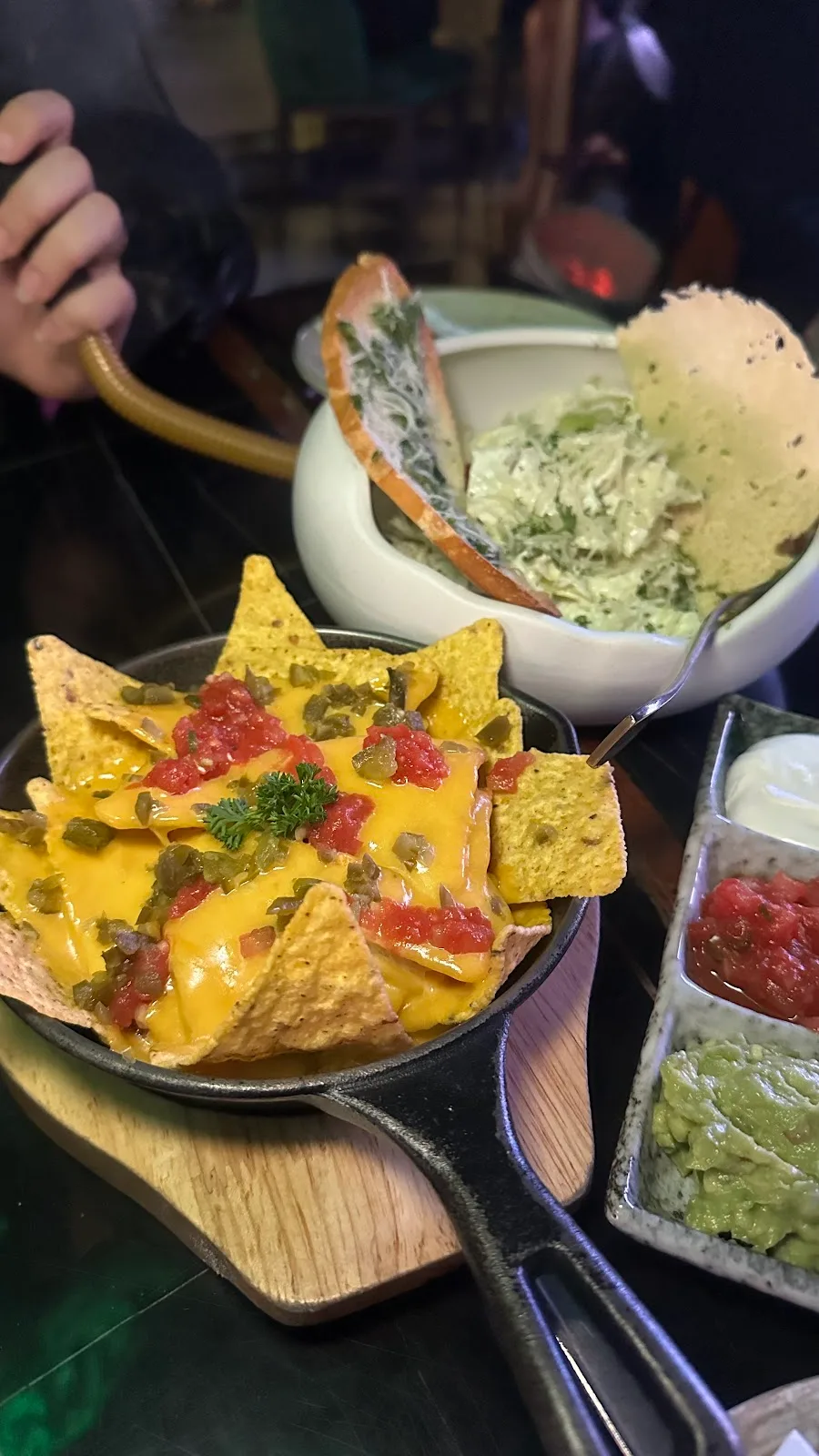 A skillet of loaded nachos with melted cheese, salsa and jalapeños beside dips of guacamole and sour cream