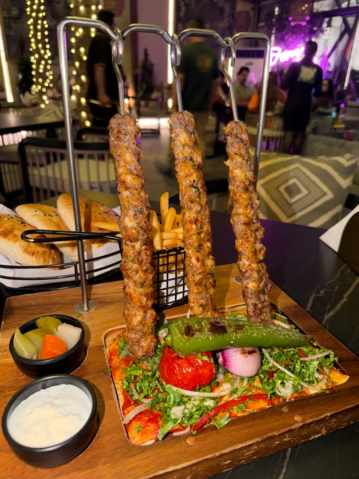 Three kebab skewers presented hanging from a metal stand over a board of salad and bread