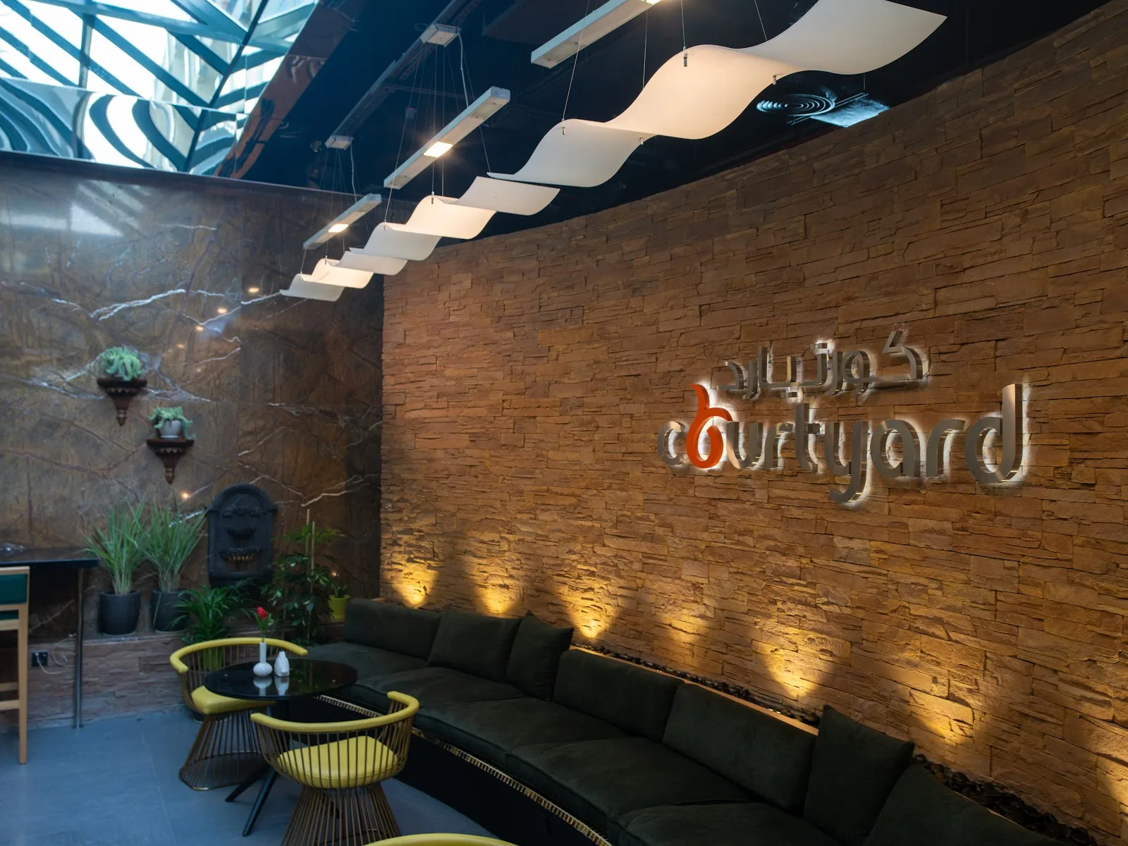 A curved stone feature wall with the 'Courtyard' sign, a long green sofa and sculptural wavy pendant lights