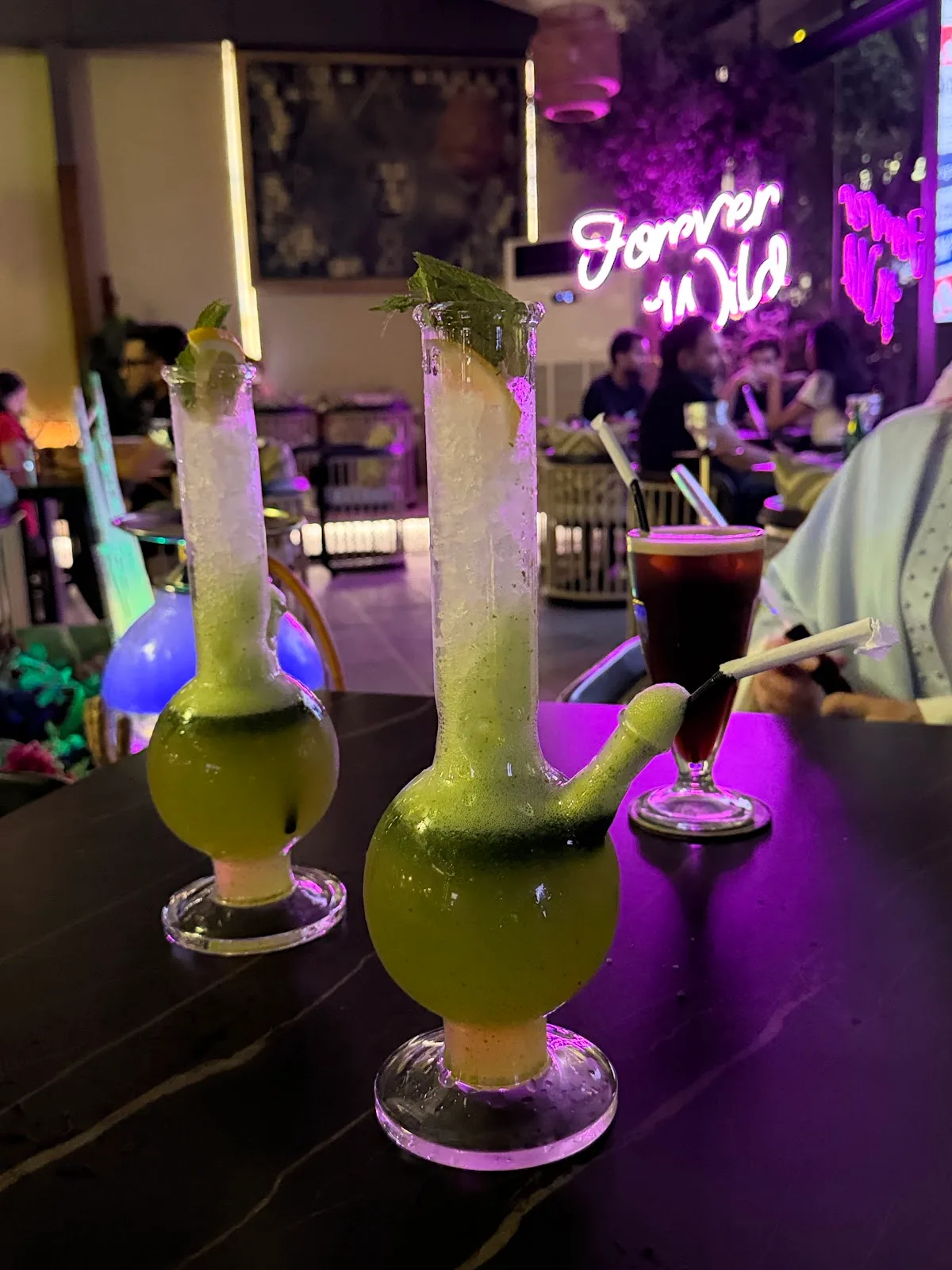 Two tall bulb-shaped glasses of bright green mint drinks on a table beneath the pink 'Forever Wild' neon sign