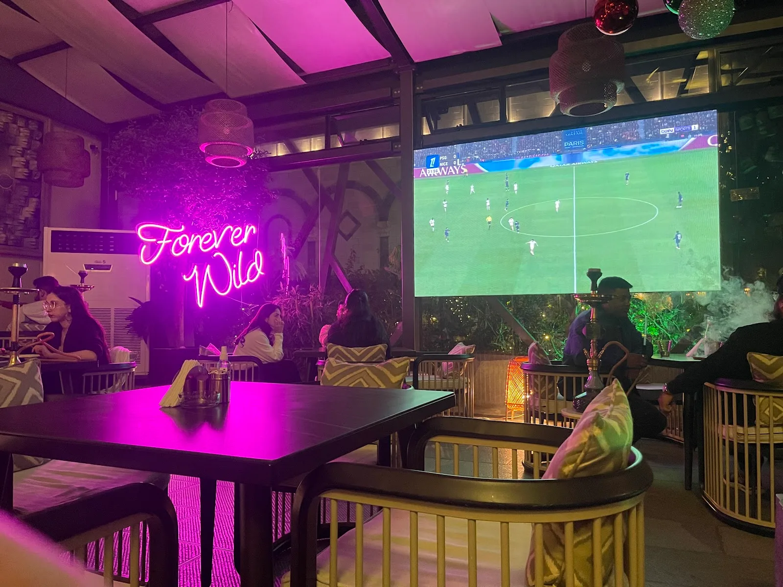 The lounge bathed in pink neon with a large screen showing live football and shisha on the tables