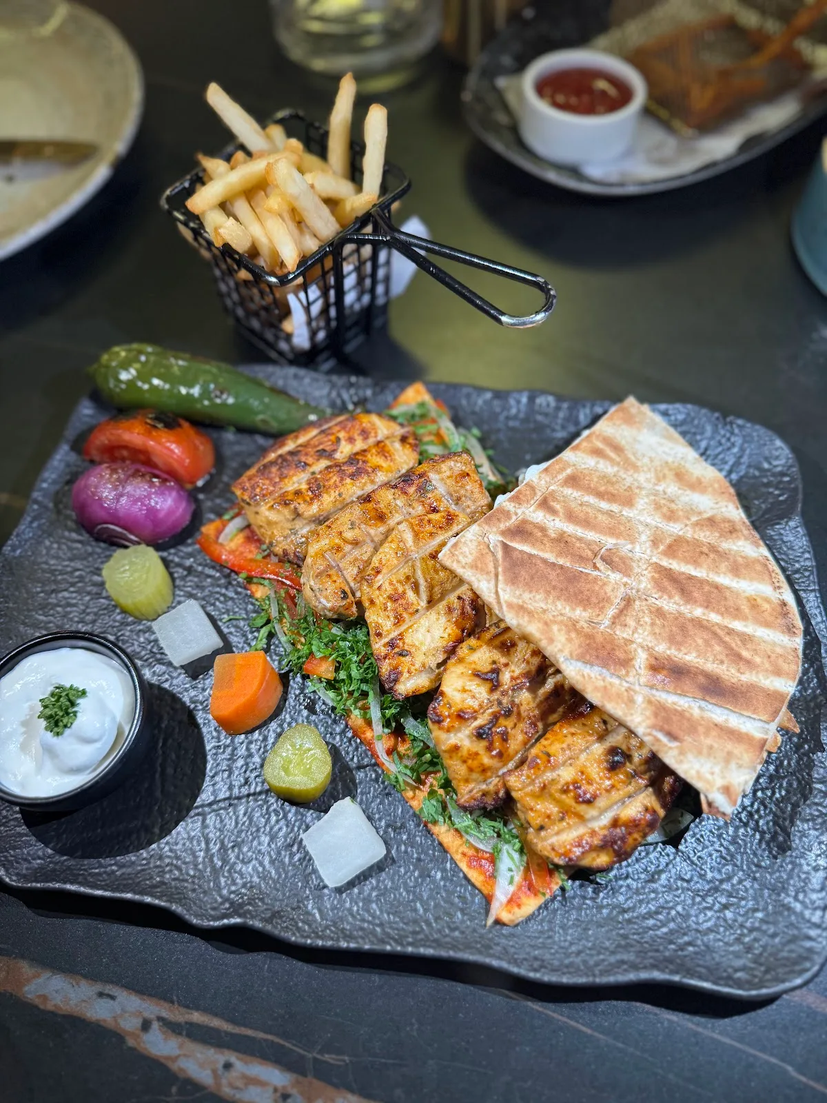 Sliced grilled chicken with grilled saj bread, pickles and a basket of fries on a slate plate