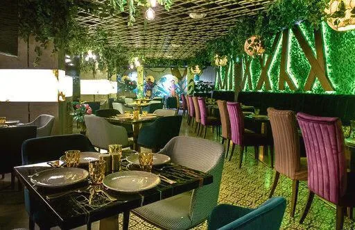 A lush indoor dining area draped with hanging vines and greenery, with jewel-toned velvet chairs and gold tableware