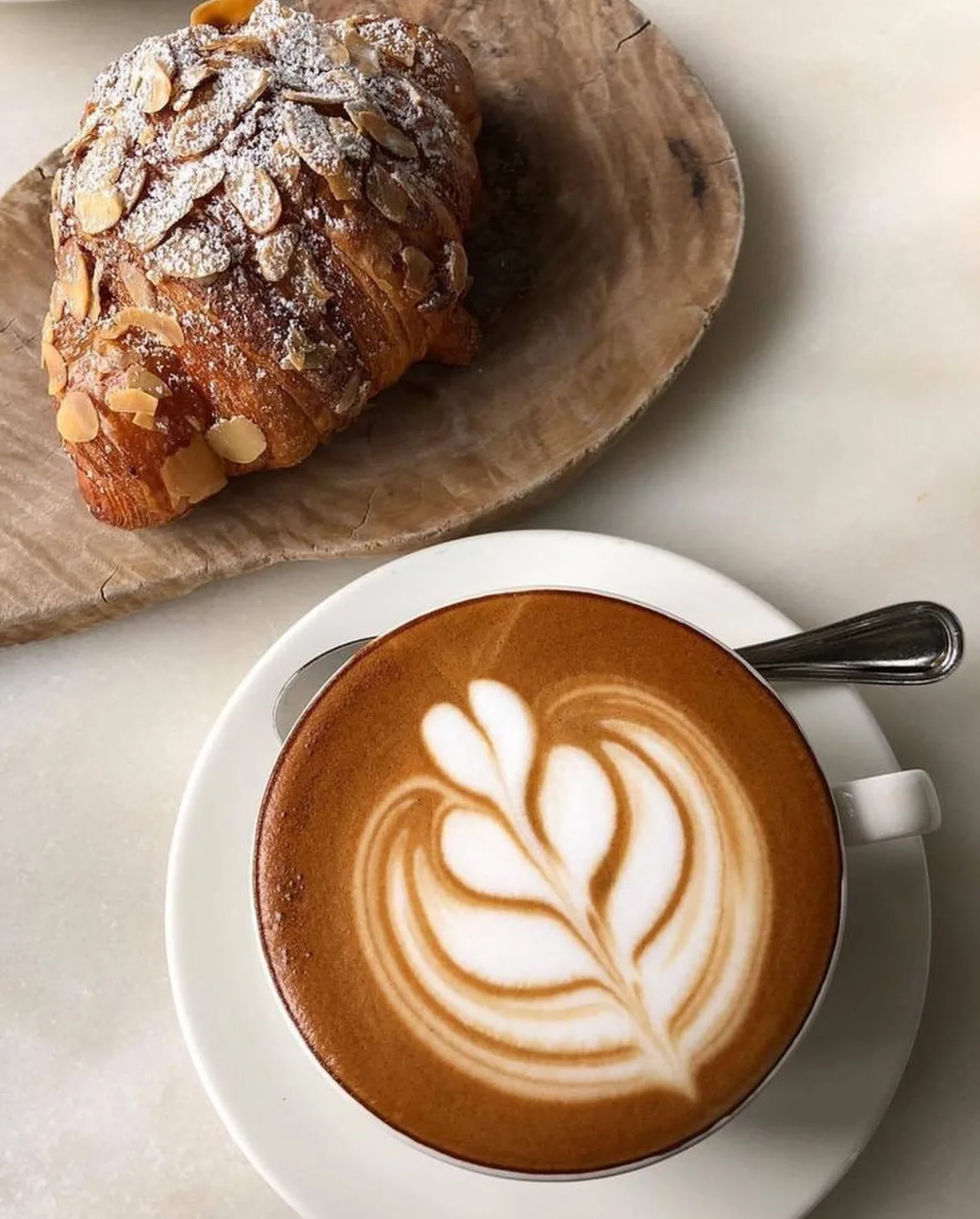 A flat white with leaf latte art beside an almond croissant on a marble café table