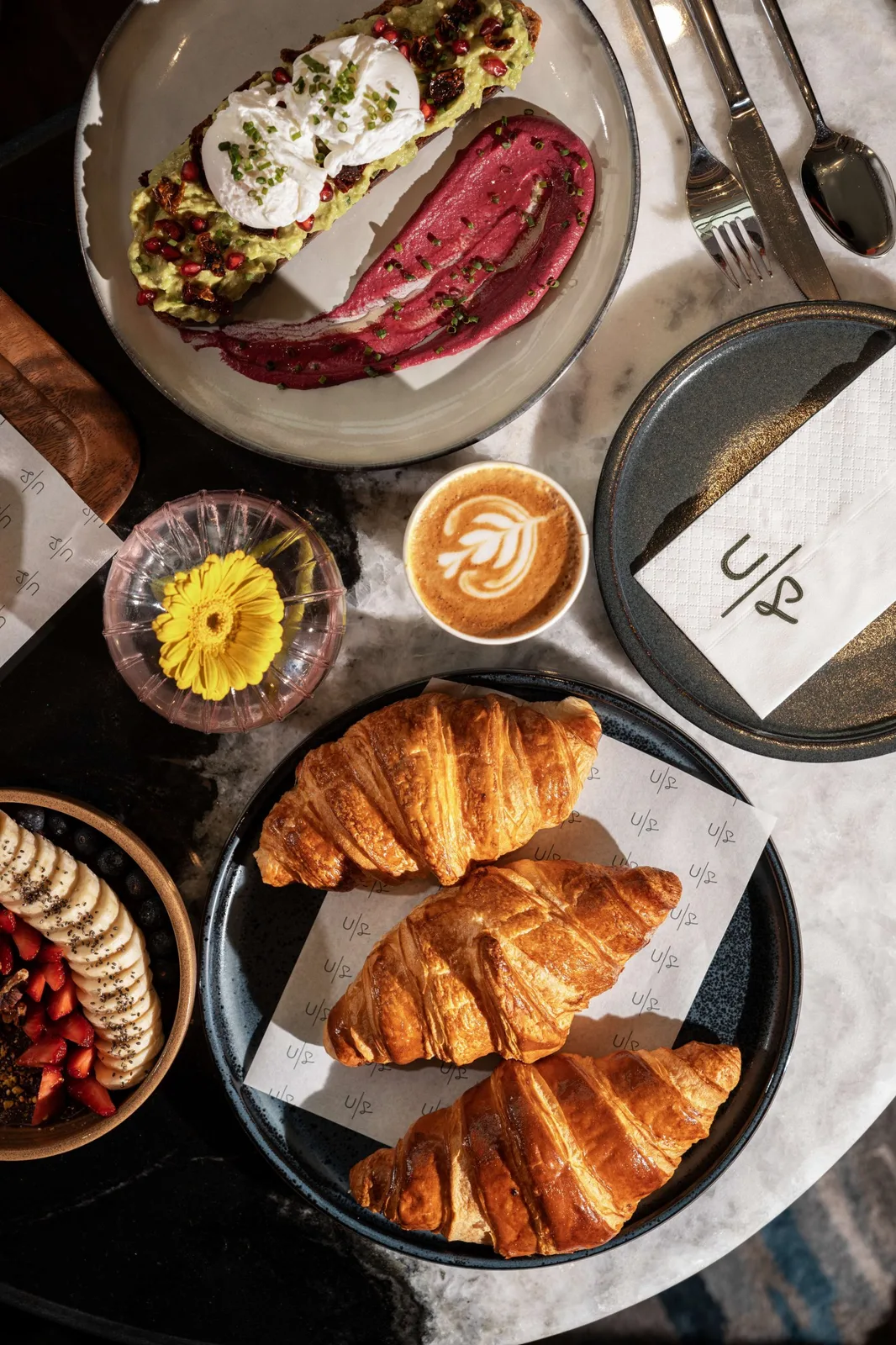 Overhead flat-lay of avocado toast, croissants, açaí and a branded flat white