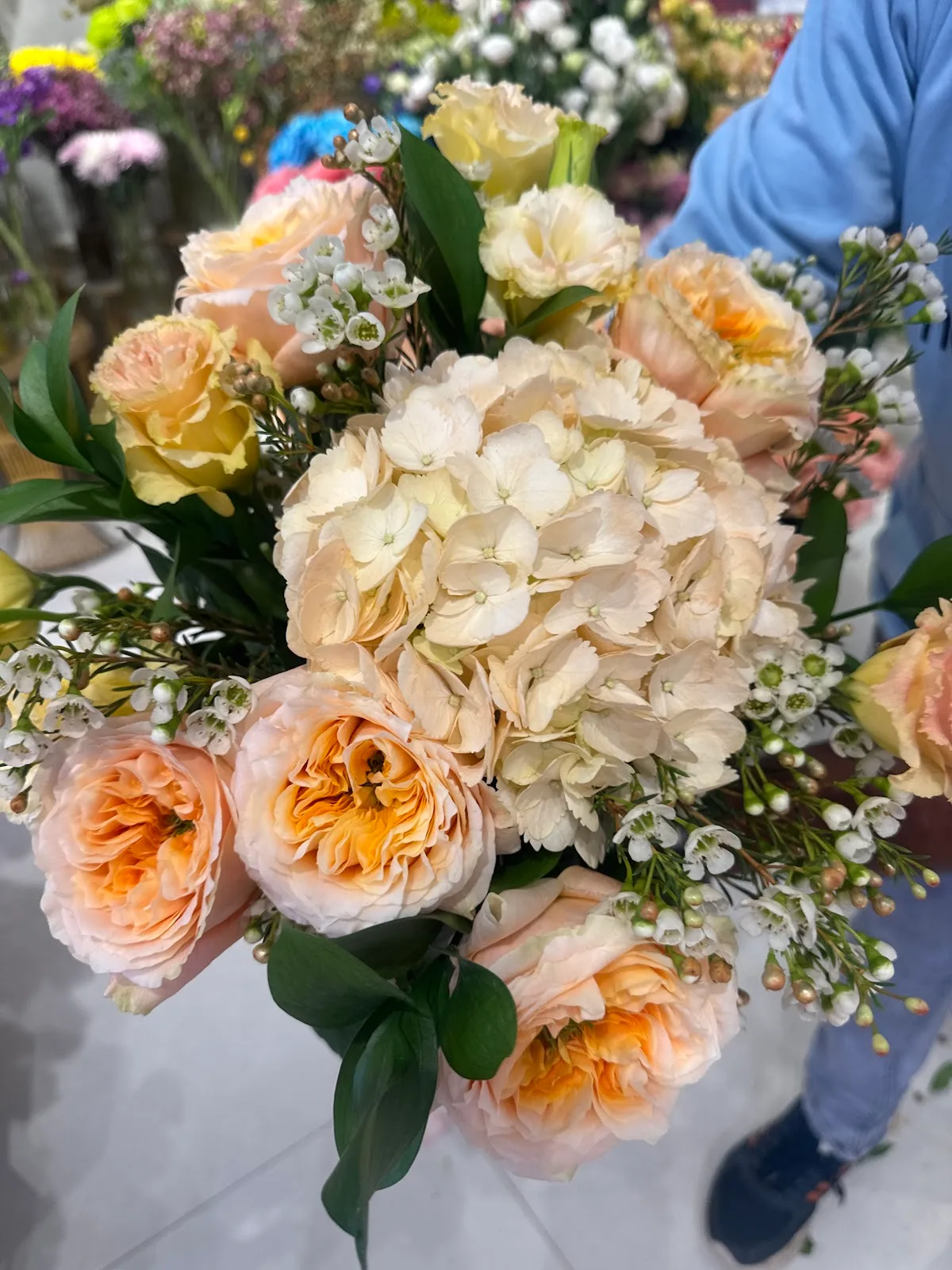 A hand-tied bouquet of peach garden roses, cream hydrangea and wax flower, held in the shop.