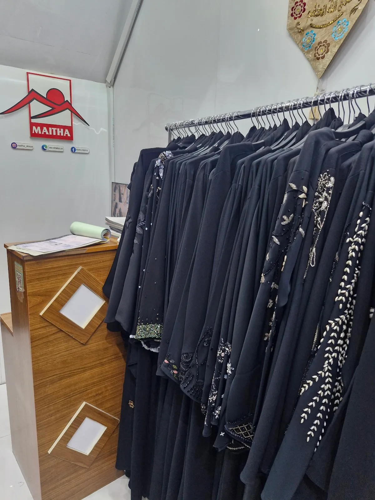 Rack of beaded black abayas beside the red Maitha logo inside the Deira shop