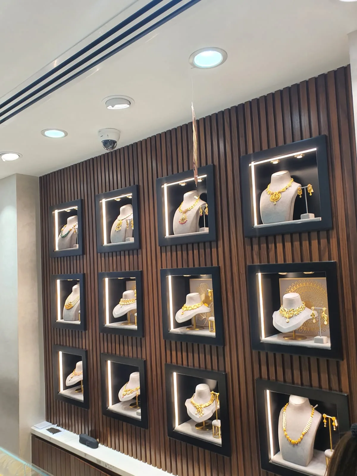 Lit black display frames on a wood-slat wall, each holding gold necklace sets at Thangals