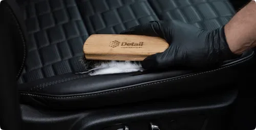 A gloved hand brushing and cleaning a leather car seat with a Detail-branded brush