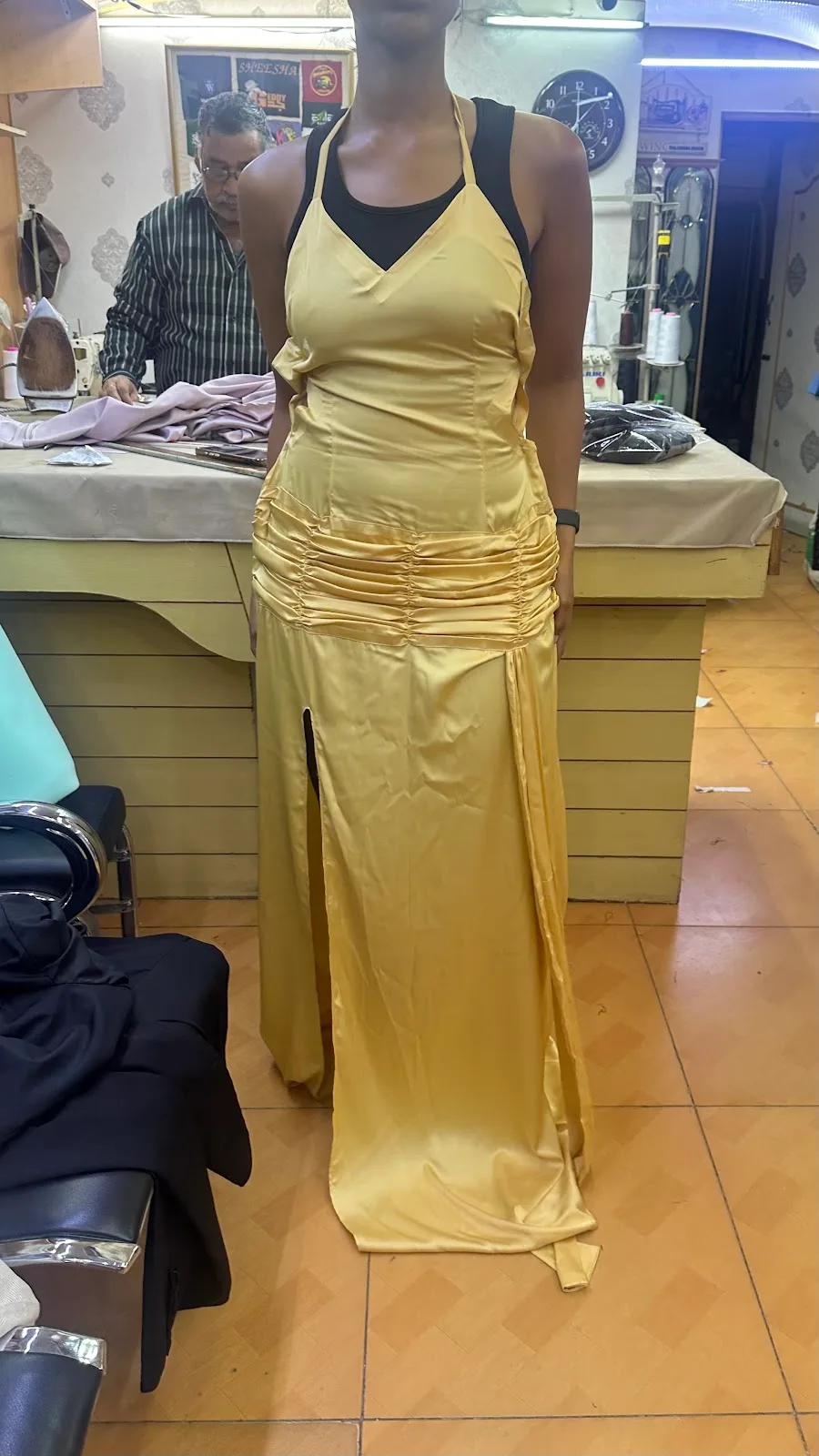 A custom gold satin evening gown fitted on a client at Electra Tailoring, with a tailor working at a sewing machine behind.