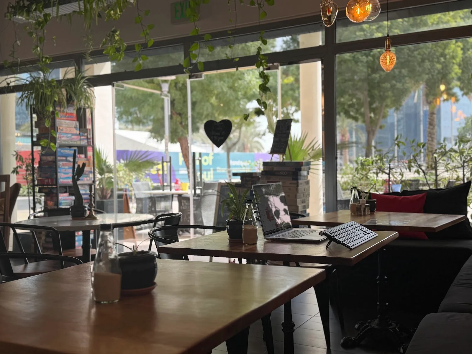 Plant-filled café interior with wooden tables, hanging bulbs and a games shelf by the window