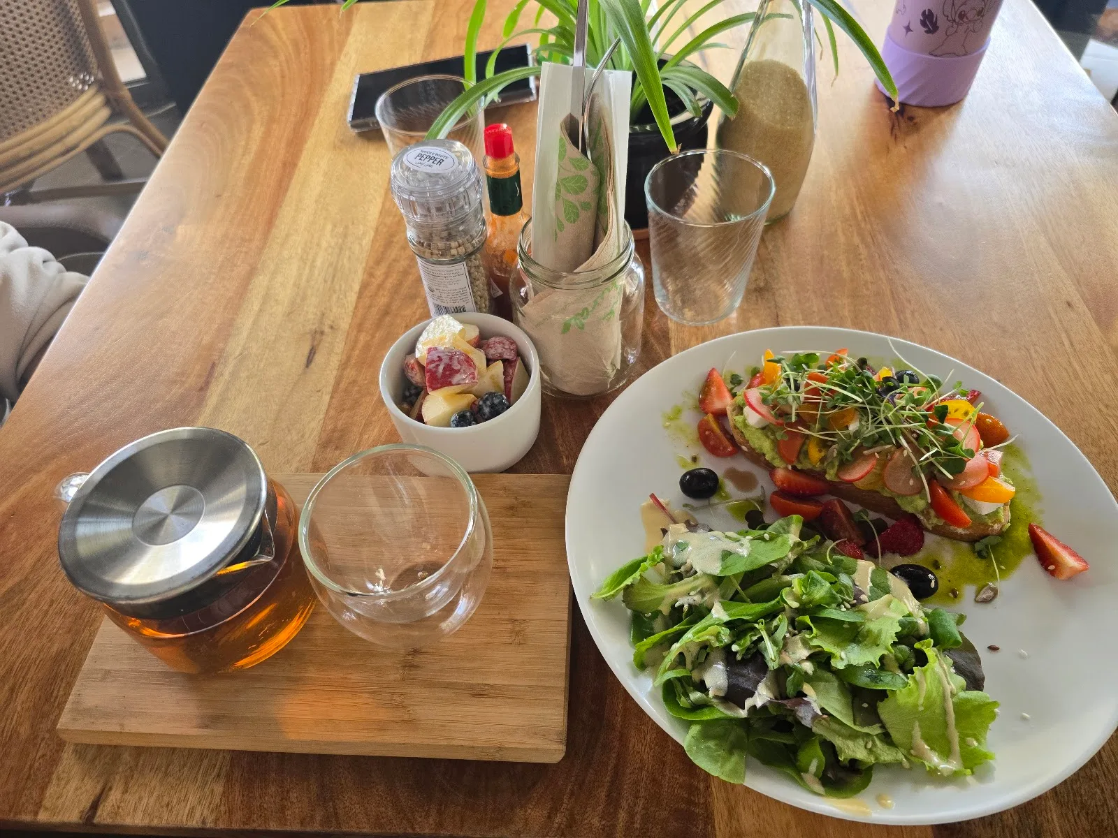 Brunch spread with avocado toast, a leafy salad, fruit bowl and a pot of tea on a wooden table