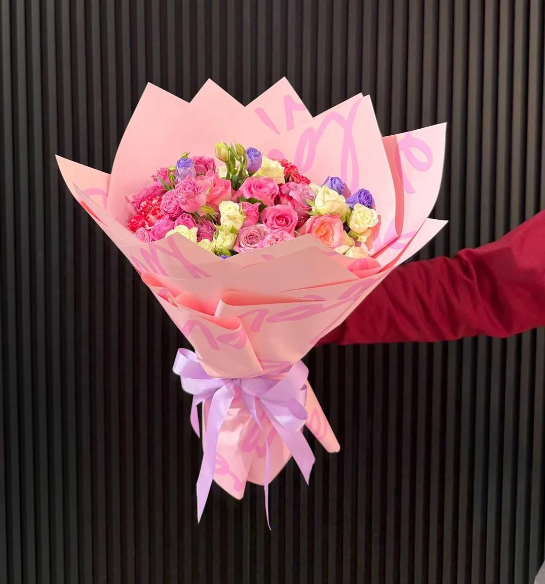 A pink-wrapped bouquet of mixed garden roses held against a dark fluted wall