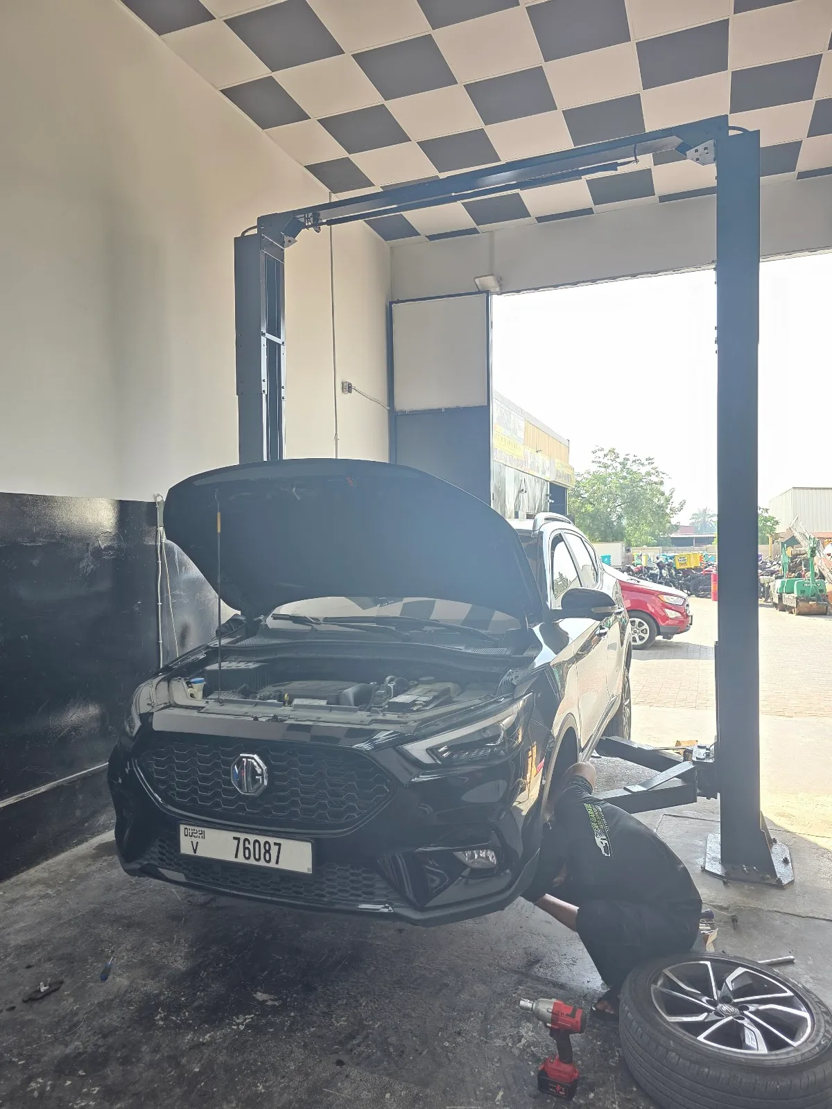 A car raised on a two-post lift inside the workshop with the bonnet open and a technician at the wheel