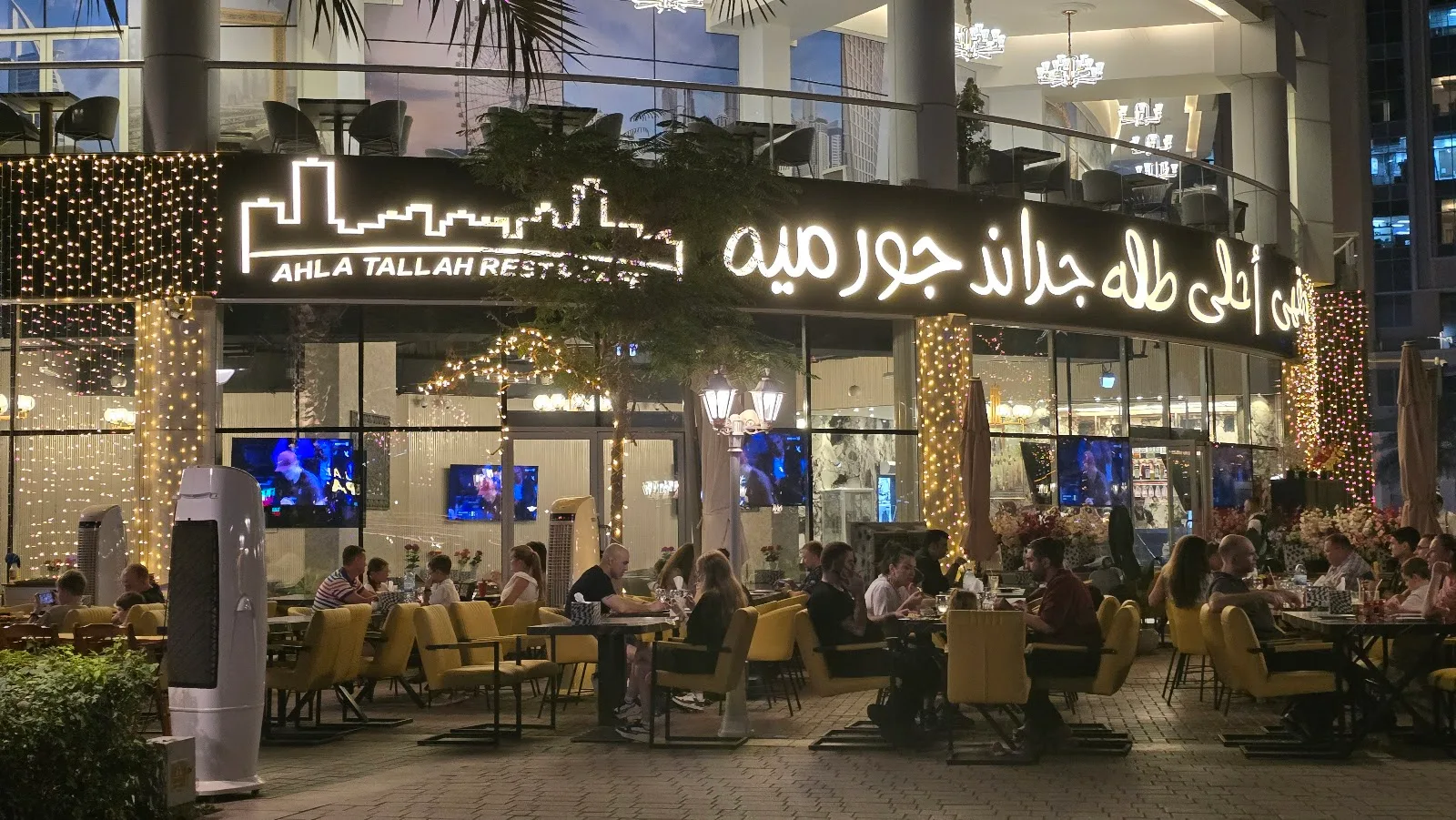 The illuminated Ahla Tallah Restaurant frontage at night, with its skyline-style sign and fairy-lit trees over the terrace