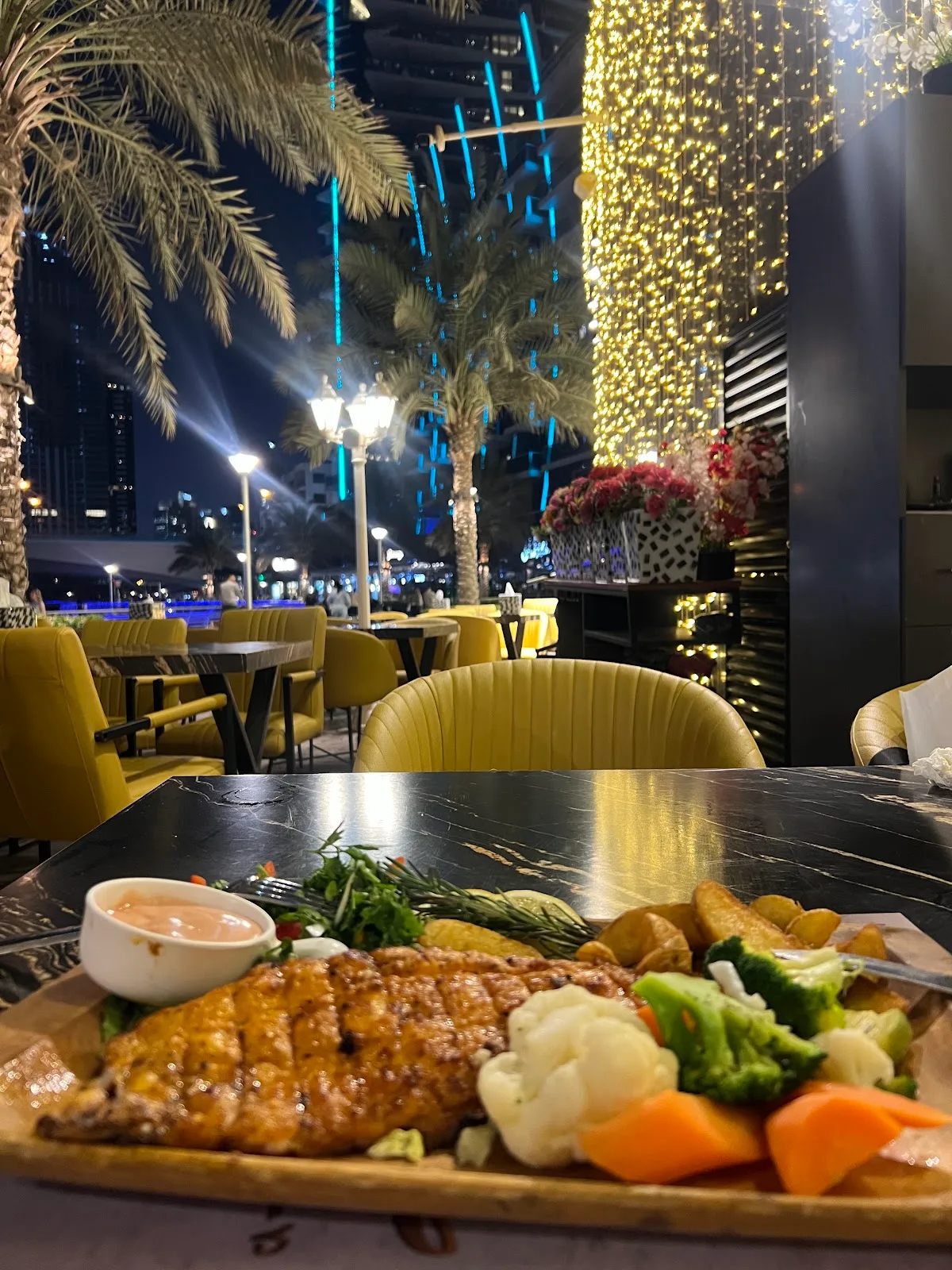 Grilled main course on a marble table on the terrace, with Dubai Marina towers and fairy lights glowing behind