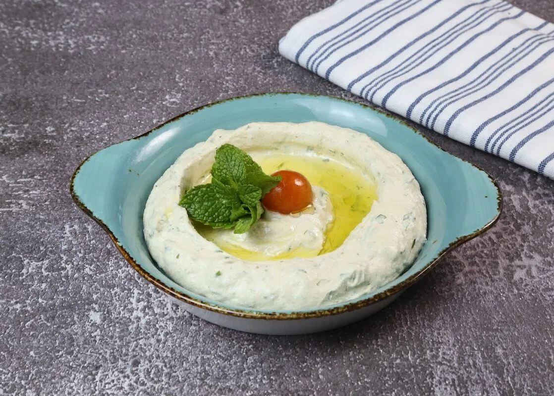 A bowl of hummus topped with olive oil, herbs and a cherry tomato on a dark stone table