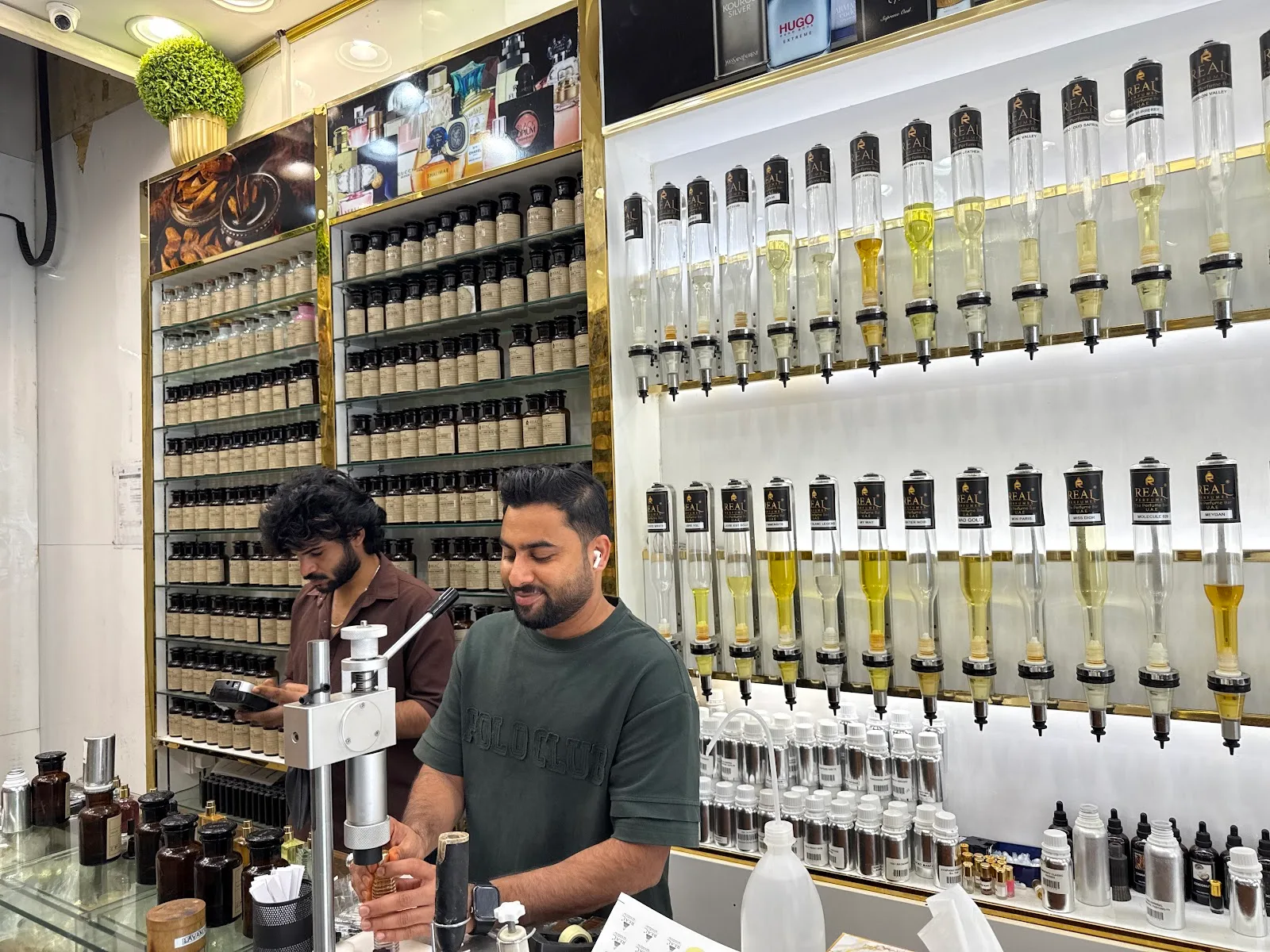 Two perfumers at the Real Perfumes counter beside shelves of raw materials and rows of fragrance oil dispensers