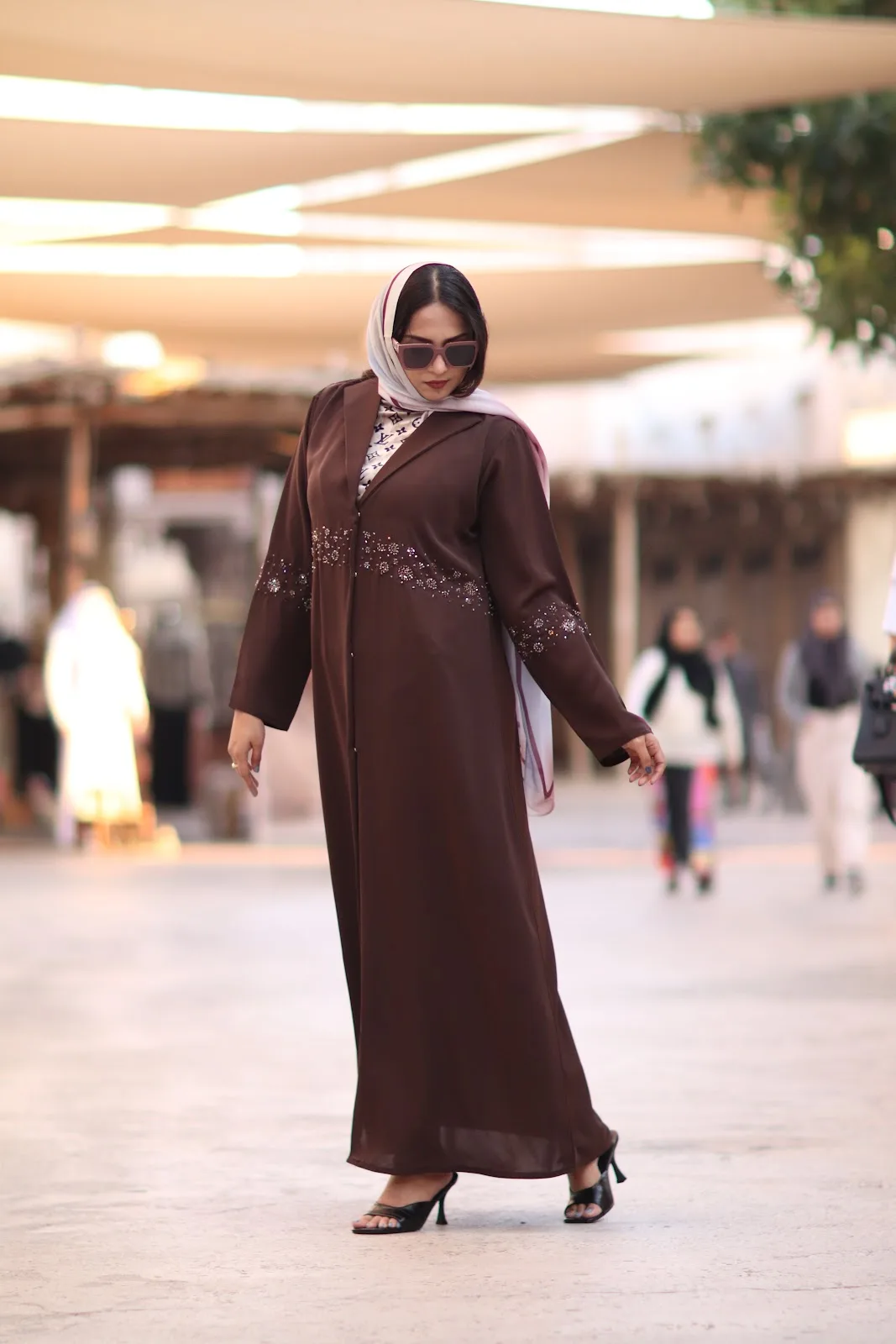 Model in a brown embellished abaya walking through a sunlit souk