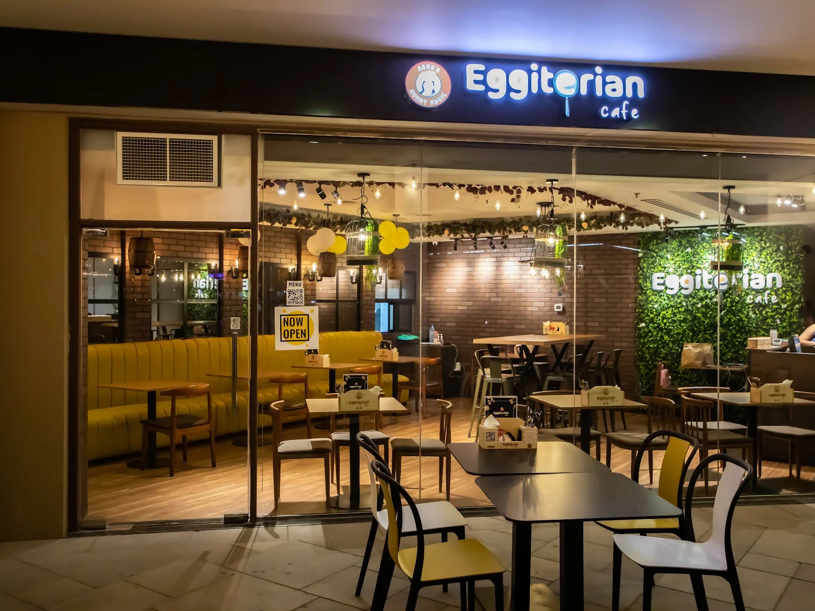 Eggiterian Cafe storefront at night in Jumeirah Lakes Towers, with its glowing wordmark sign, green living wall and warm yellow seating