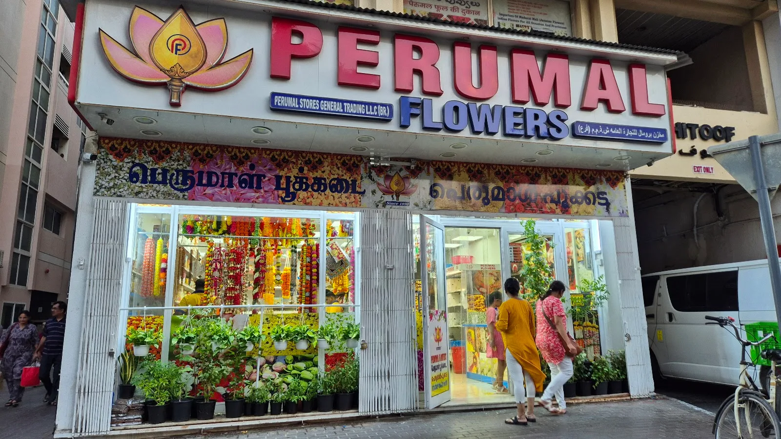 The Perumal Flowers storefront in Al Karama, Dubai — a glass shopfront filled with fresh garlands, potted plants and loose flowers under the red and blue PERUMAL FLOWERS sign with its pink lotus mark.