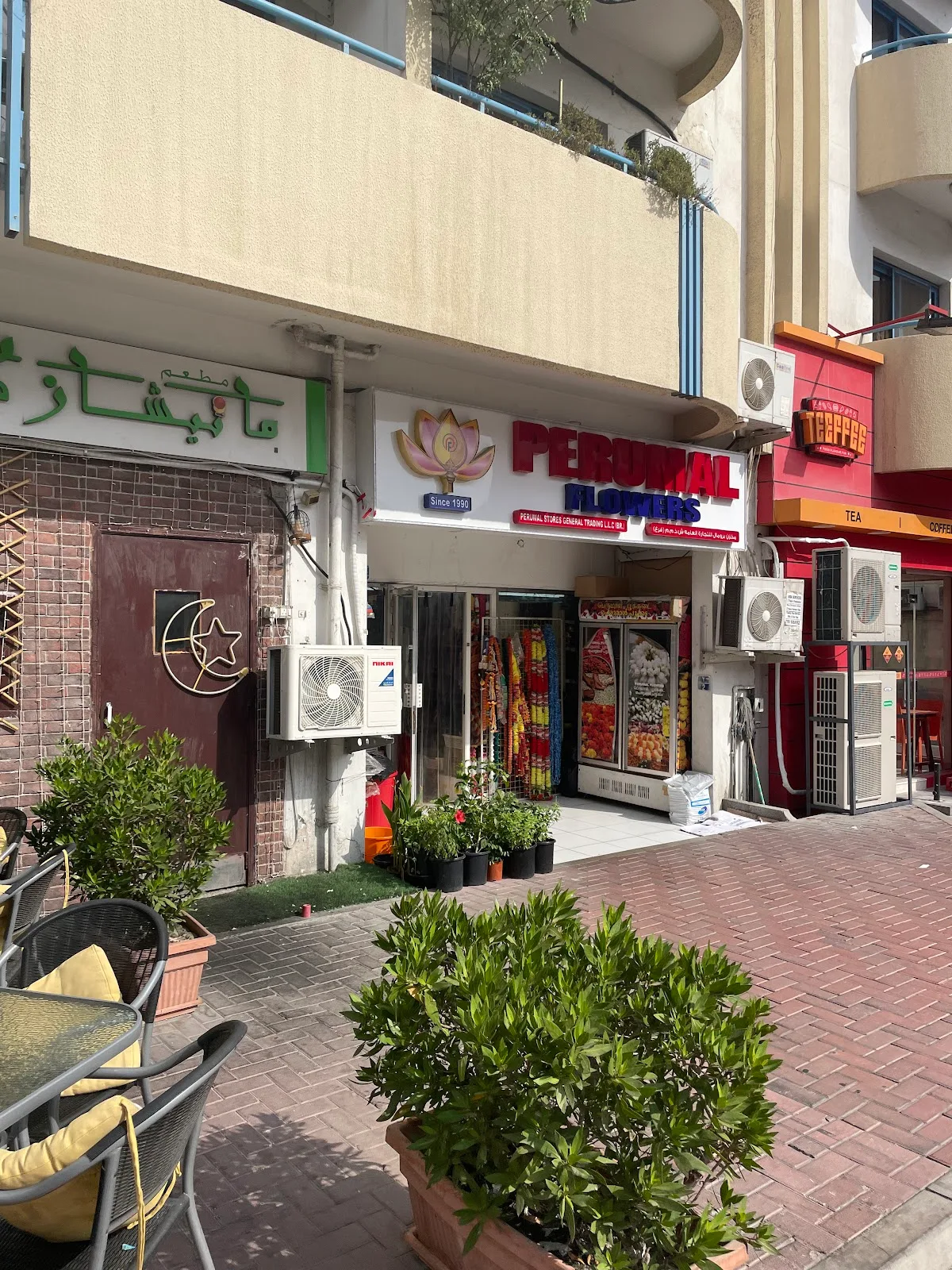The Perumal Flowers shopfront with its 'Since 1990' lotus sign on a quiet Karama street, potted plants outside and a café nearby.